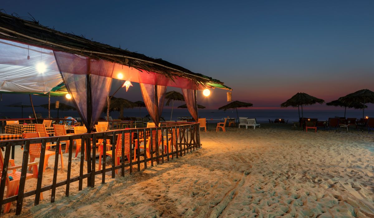 A cozy wooden beach shack in Goa lit with warm orange lanterns at dusk, featuring orange chairs on the sand, perfect for a romantic dinner on our goa holiday package 2 nights 3 days