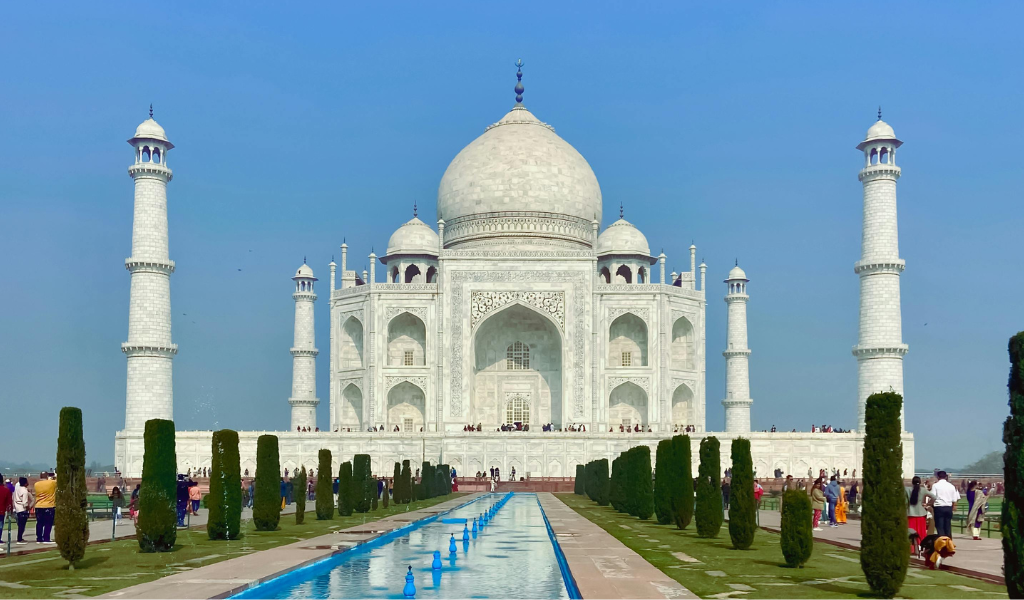 The majestic white marble Taj Mahal in Agra with reflection pool and green trees under a blue sky, a key highlight of our Golden Triangle with Goa tour package