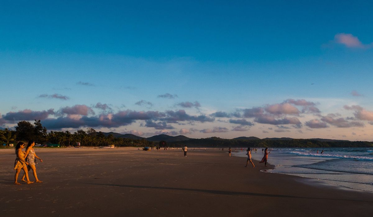 Two travelers walking along a wide, sandy Goa beach during the golden hour with mountains in the background, a top experience included in our 2 nights 3 Days Goa Tour