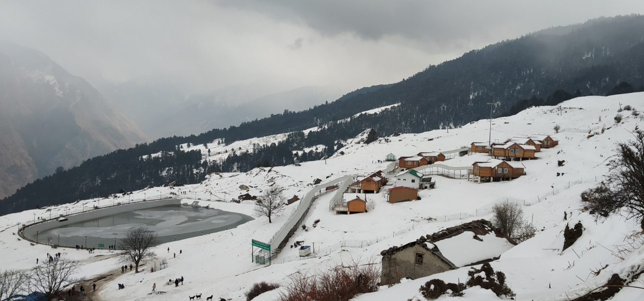 Auli tour package showing snow-covered artificial lake and wooden cottages set against winter mountain slopes.