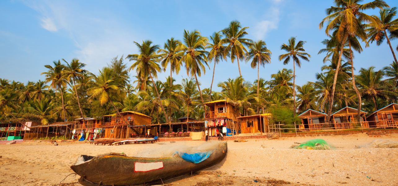 A traditional Goan fishing boat rests on the sand in front of vibrant, colorful beach shacks and tall palm trees, a unique cultural experience included in couples 2026 Goa holiday tour package.