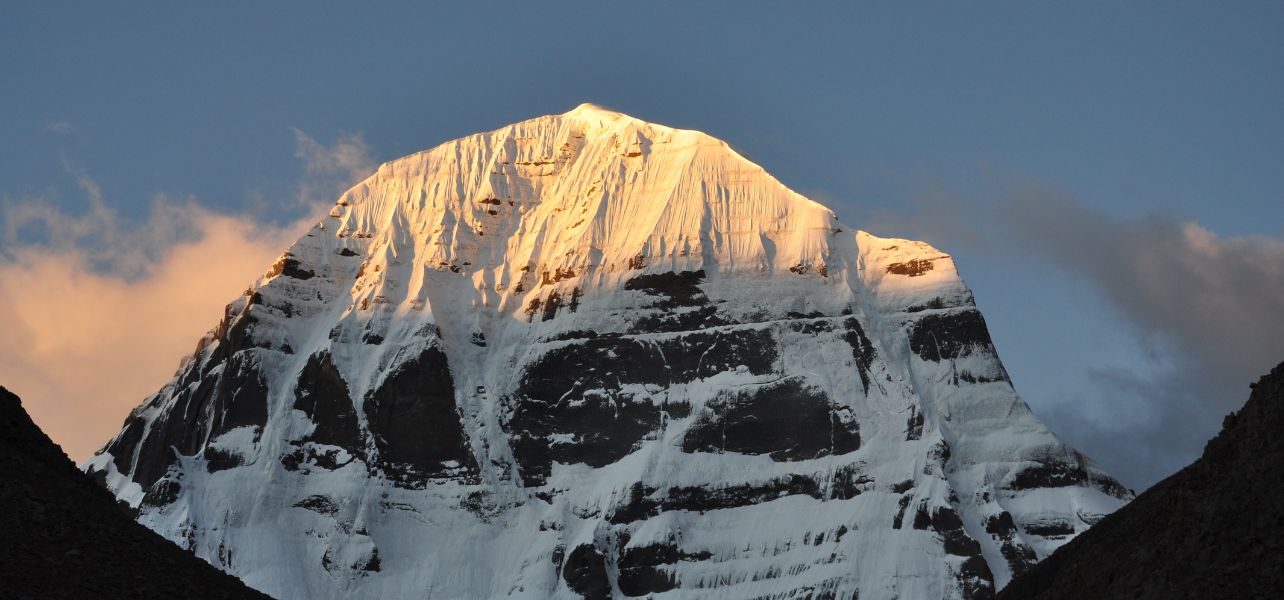 In The image Golden sunlight hitting the snow-covered peak of Mount Kailash During the Trip To Kailash Mansarovar against a clear blue sky during sunset.