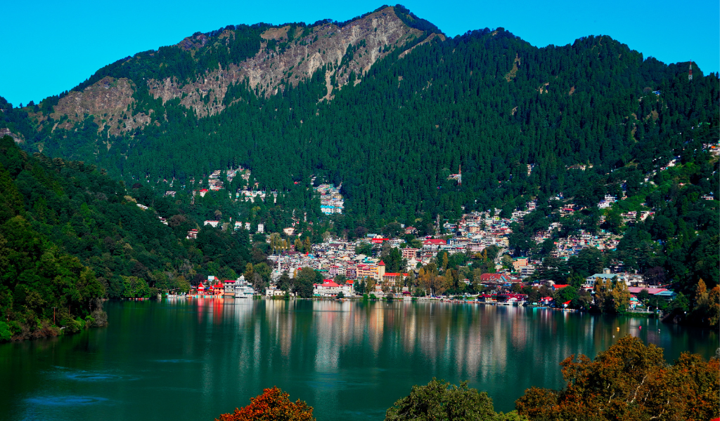 A stunning panoramic view of the Naini Lake in Nainital, surrounded by lush green mountains and colorful houses, as included in our Nainital Corbett With Valley of Flower tour package.