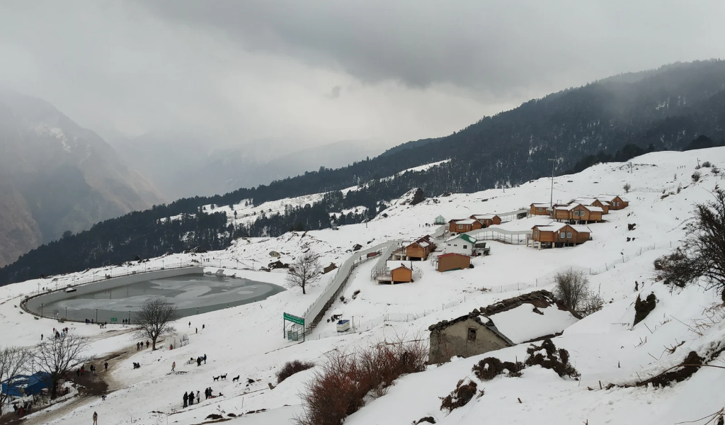 Snow covered landscape view of Auli artificial lake and wooden cottages included in Auli Nainital tour package.