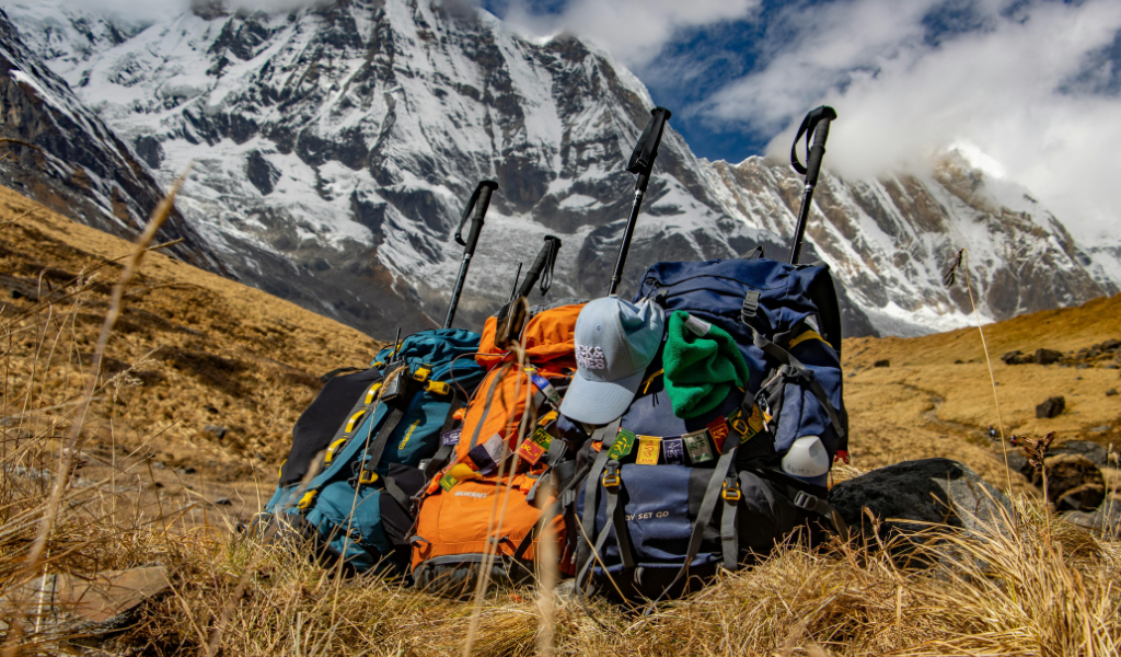 Image of Trekking backpacks, trekking poles, and caps resting on alpine meadows with snow-capped mountains in the background captured during the Kuari Pass trek.