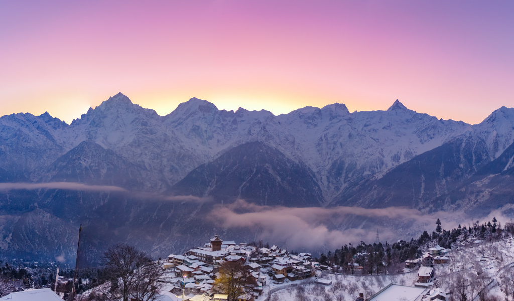 A beautiful village covered in white snow with high mountains and a purple and pink sunset sky in the background.