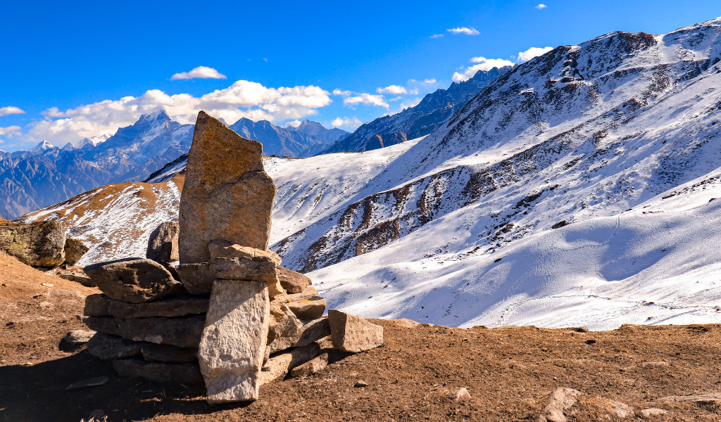 Stone cairn landmark on the Kuari Pass trek package trail with snow-covered Himalayan mountain peaks under clear blue sky.